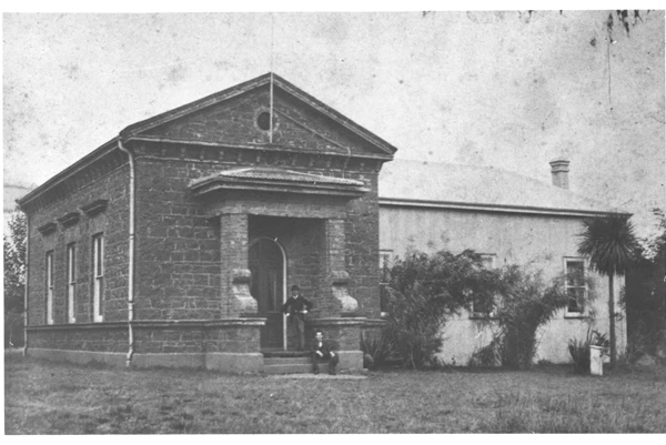 A black and white photo of a grey, stone building on a sparse lawn. A square archway leads to a door at the front. Two people pose on the step under the archway. The building continues to the right with a few trees obscuring several windows.