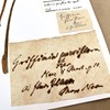 Several labels are affixed to the corner of a white piece of card. The largest is pale brown and mottled, with cursive handwriting in ink which has bled into the paper. Next to the label are two pressed, brown stems of a specimen. 